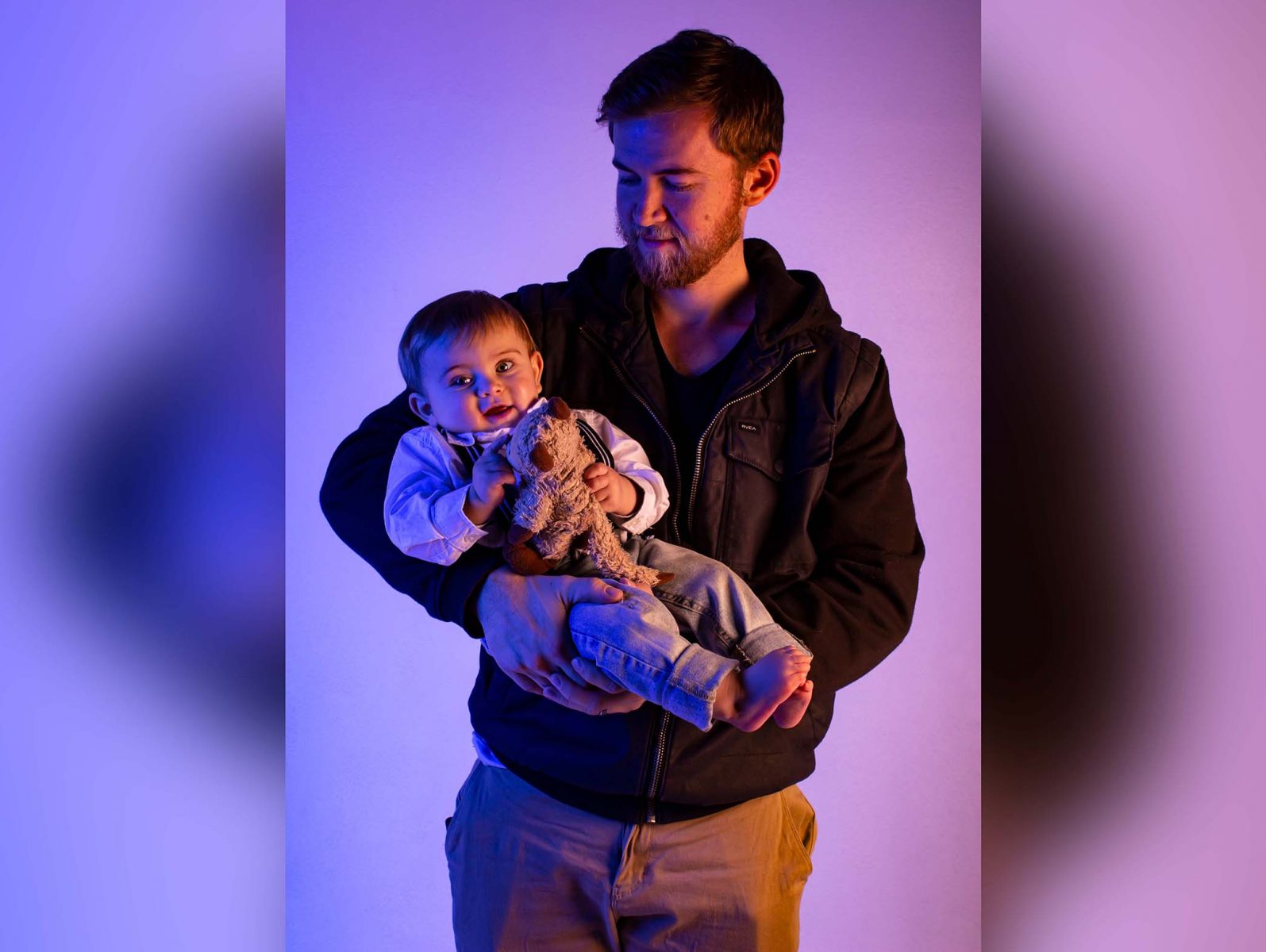 Father holding a 1 year old agains a purple and pink background